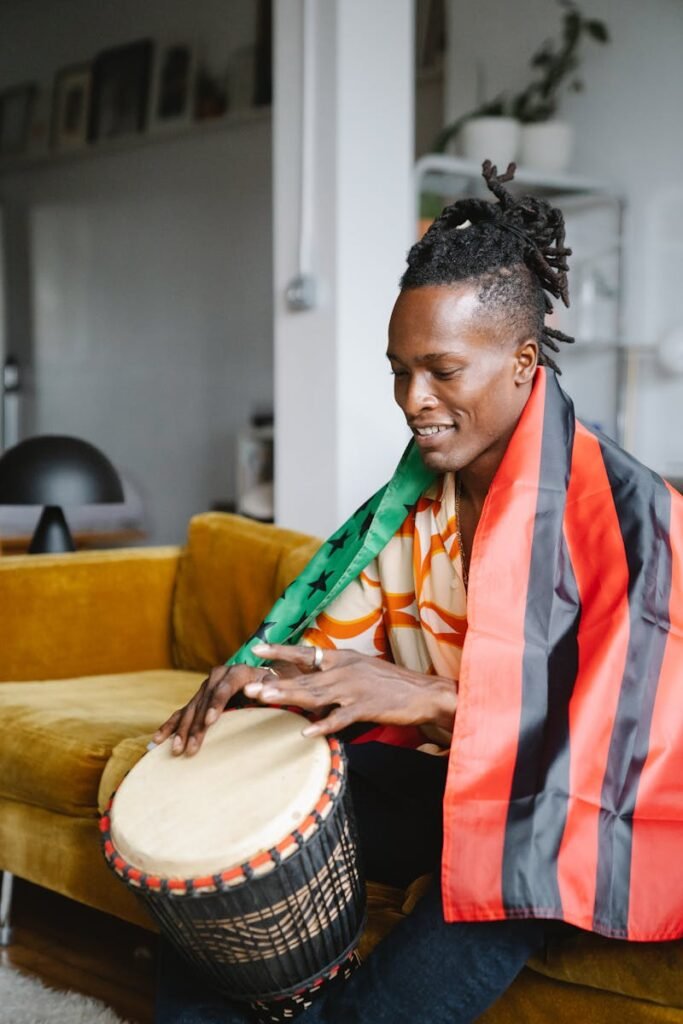 African musician with dreadlocks plays djembe drum indoors wrapped in a colorful flag.