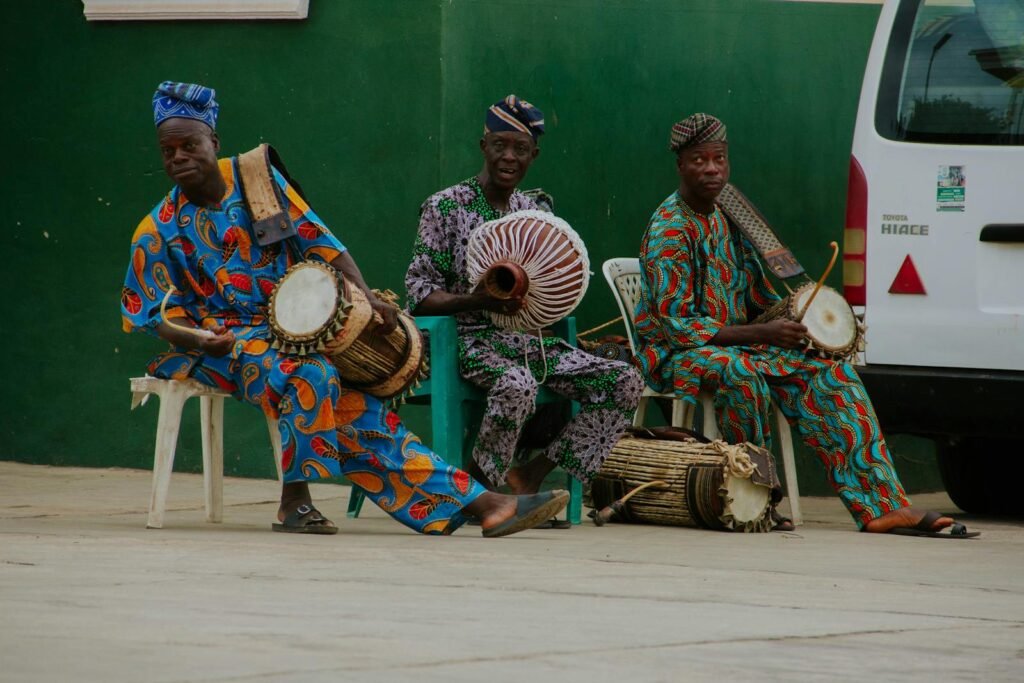 Three African men play traditional Yoruba drums in vibrant attire outdoors in Ogbomoso, Nigeria.